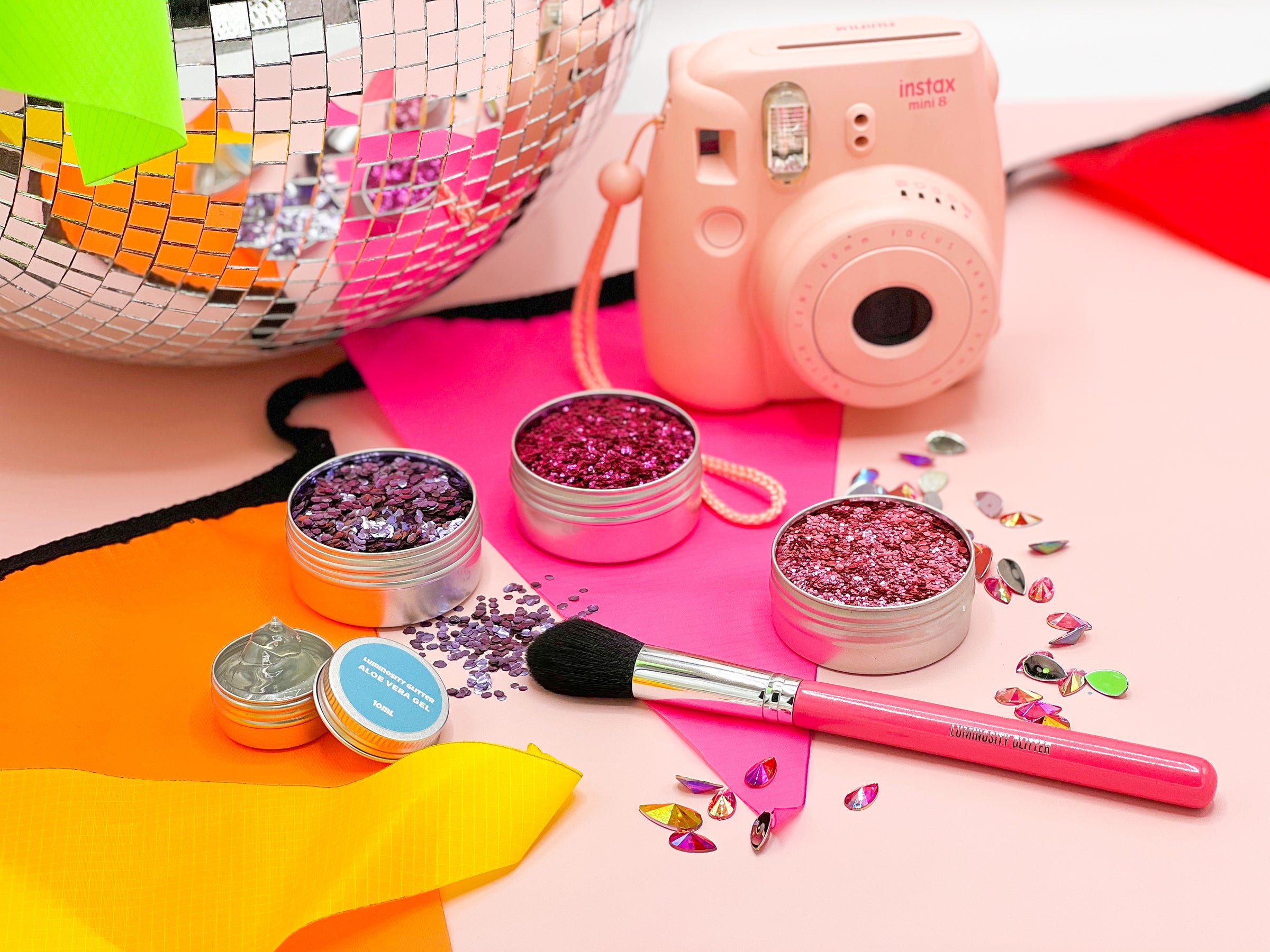 Neon bunting, a pink Polaroid camera and giant disco ball with eco glitter and a hot pink makeup brush. Sprinkled on a pink background are reusable face gems