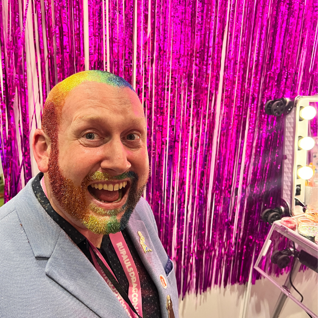 A bald man with a glitter rainbow head and glitter rainbow beard from Drag Con UK 2023