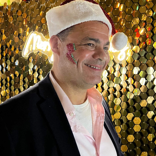 Gold luxe sequin wall with a gentleman standing infront wearing a santa hat with a pink and green glitter lightning bolt on the side of his face.